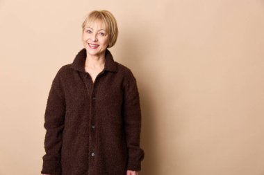 Kısa saçlı, koyu kahverengi ceketli mutlu kadın bej arka planda poz veriyor. Günlük kıyafet, mevsimlik kıyafet, doğal görünüş ve kendine güvenen ifade anlayışı..