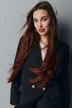Gri duvarın yanında duran siyah ceketli, dalgalı kahverengi saçlı, kendinden emin genç bir kadın. Doğal görünümlü bir kadın portresi ve yaşam tarzı, moda veya güzellik teması için günlük kıyafetler..