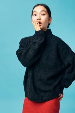 Siyah örgü süveter ve kırmızı etek giyen genç bir kadın ağzının yanındaki elle şok olmuş görünüyor. Moda ve duygusal tepkiler stüdyoda mavi arka planda çekildi.