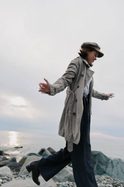 Paltolu ve şapkalı genç bir kadın okyanus yakınlarındaki bulutlu gökyüzünün altında kayalık sahilde yürüyor. Sonbahar sezonu boyunca rahat bir yaşam tarzı, günlük moda, denge ve doğa bağlantısı..