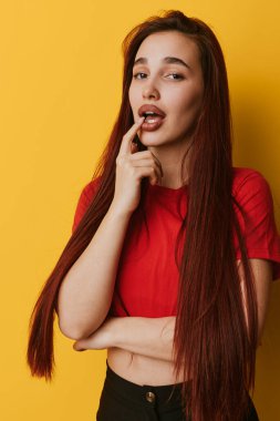 Sarı arka plana karşı kırmızı saçlı, uzun saçlı genç bir kadının portresi. Dudakları üzerinde parmak pozu, rahat duruş ve doğal görünüm yaşam tarzı, moda veya güzellik konsepti.