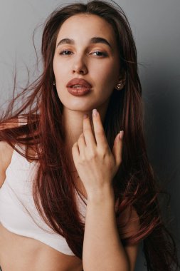 Dalgalı saçlı, doğal görünümlü, çenesine dokunan, sıradan beyaz elbiseler giyen ve arka planda sade ışıkta duran küçük küpeli genç bir kadının portresi..