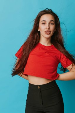 Kendine güvenen, uzun saçlı, kırmızı kısa kollu üst ve mavi arka planda duran siyah, yüksek bel pantolonlu bir manken. Sıradan moda ve rahat ifade ile portre.