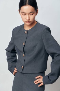 Dekoratif düğmeli gri şık takım giyen Asyalı kadın modelin portresi. Minimalist stüdyo ortamında kadın elleri belinde poz veriyor. Yumuşak ışıklandırma ve tarafsız zemin ile..