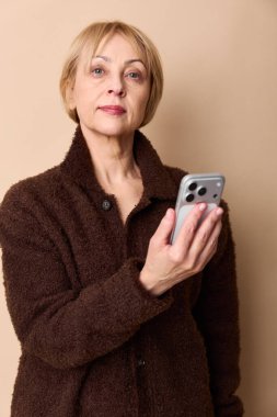 Bej duvarda duran kahverengi yün kazak giyen orta yaşlı kadın portresi. Üst düzey teknoloji kullanımı, iletişim ve dijital bağlantı kavramı.