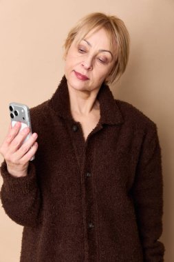 Kısa saçlı, kahverengi ceketli, elinde akıllı telefon olan ve bej arka planda dikkatlice ekrana bakan bir kadın. Dijital bağlantı, teknoloji kullanımı, gündelik moda ve yaş çeşitliliği kavramı.