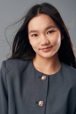 Gri ceketli, uzun siyah saçlı, gülümseyen genç bir kadının portresi. Stüdyoda duruyor. Güzellik, moda ve sıradan görünüş için az bir geçmişi var..