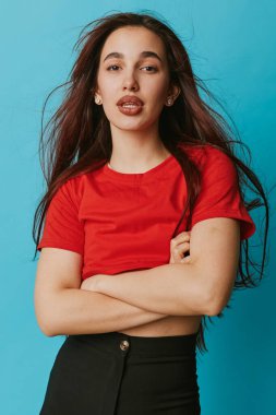 Uzun saçlı, kırmızı tişörtlü, siyah pantolonlu ve kolları mavi arka planda çapraz duran kendine güvenen bir kadın portresi. Sıradan moda, doğal görünüş ve stüdyo ortamında rahat duruş..