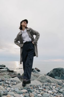 Bulutlu bir günde çakıl taşlı sahilde paltolu ve şapkalı kendine güvenen genç bir kadın poz veriyor. Moda, doğa, açık hava yaşam tarzı, sakin ve doğal ortamda yakalanan sıradan tarz..