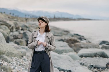 Eski moda kıyafetler giymiş şık genç bir kadın sakin suların kıyısında kayalık sahilde poz veriyor. Dokulu palto ve şapka giyiyor, gözleri kapalı, sakin kıyı havasına dalmış, dağlar kadar uzakta..