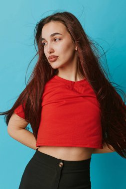 Uzun kahverengi saçlı, kırmızı bluzlu, mavi pantolonlu, rahat duruş ve doğal makyajla yüzünü çeviren kendine güvenen genç bir kadının portresi..