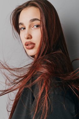 Uçan kızıl saçlı ve kameraya bakan makyajsız genç bir kadın. Yumuşak ışıklandırmalı yakın plan portresi ve nötr gri arka planda günlük siyah kıyafet.