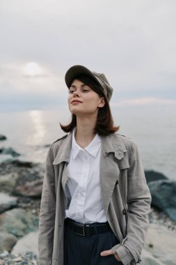 Sakin genç bir kadın, günlük palto ve kep giymiş deniz kenarındaki kayalık kıyı şeridinde, gözleri kapalı, huzurlu bir esinti hissediyordu. Rahatlama, doğa, açık hava yaşam tarzı ve farkındalık kavramı.