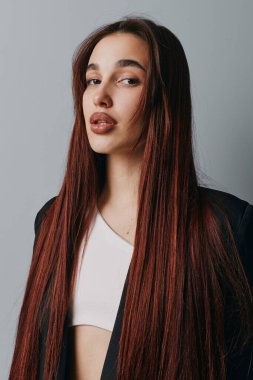 Uzun saçlı, beyaz bluzlu ve gri arka planda duran siyah ceketli genç bir kadın. Makyajlı, sakin yüzlü ve kameraya bakan kadın modelin yakın plan portresi..