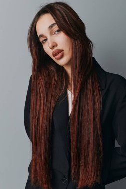 Uzun kahverengi saçlı, zarif makyajlı, siyah ceketli genç bir kadın modern tarz ve güzellik fotoğrafçılığı için gri duvarın önünde duruyor..