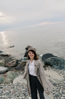 Gündelik giysiler içinde gülümseyen genç bir kadın bulutlu gökyüzünün altında sakin deniz kıyısında duruyor. Şapka takıyor ve uzun bir palto giyiyor. Açık havanın tadını çıkarıyor. Suyun yanında rahat ve mutlu bir havası var..