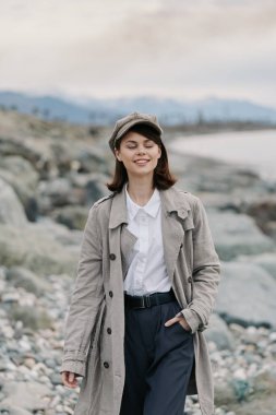 Uzun paltolu ve şapkalı gülümseyen genç bir kadın kayalık bir sahilde gözleri kapalı, huzurlu sahil çevresini ve açık havayı kucaklıyor. Sakinlik ve özgürlük kavramı.