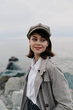 Kayalık sahilde klasik şapka ve dokulu palto giyen genç bir kadının portresi. Yumuşak gülümseme ve doğal ışık sakin bir hava yaratır. Moda, yaşam tarzı, açık hava, gündelik, klasik, doğa konsepti.