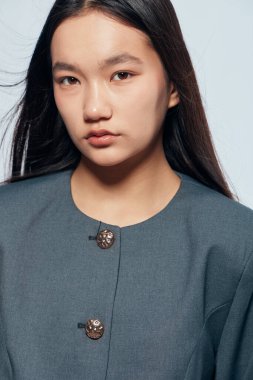 Düz siyah saçlı, dekoratif düğmeli gri bir ceket giyen Asyalı genç bir kadının portresi. Temiz bir arka plan, en az mücevher ve moda tarzında tarafsız yüz ifadesi..
