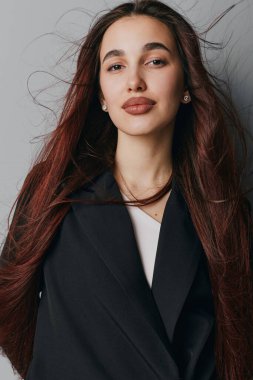 Uzun kahverengi saçlı, kısa makyajlı, kendine güveni tam bir kadın portresi. Gri arka planda poz veren küpe, doğal bir ifade ve iç mekan tarzı var..