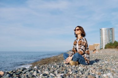 Suyun yakınındaki kayalık bir sahilde güneş gözlüğü ve kot giyen sakin bir ufuk, esinti ve güneşli bir anda deniz atmosferinin tadını çıkaran sıradan bir kadın figürü..