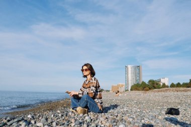 Sakin suların kıyısındaki çakıl taşlı bir sahilde gündelik gezgin, ekose gömlek ve kot giyen kadın güneşli bir günün tadını çıkarıyor şehir arkaplanı ve geniş mavi ufkuyla.