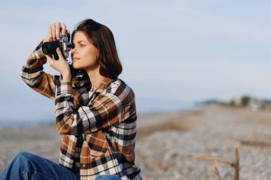 Sahildeki kadın fotoğrafçı, kamerayı denize ve ufka doğrultuyor, doğal ışık ve sakin esintiyle kıyı sahnesini çekiyor..