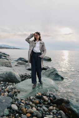 Trençkot ve koyu renk pantolon giyen genç bir kadın sakin deniz kıyısında kayalık sahilde duruyor. Yumuşak güneş ışığından korunuyor. Bulutlu gökyüzü ve sakin su, dışarıda huzurlu bir anın altını çiziyor..