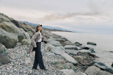 Günlük giysiler giyen genç bir kadın kayalık sahilde durup bulutlu gökyüzü ile sakin denize bakıyor. Açık hava yaşam tarzı, doğa keşfi, rahatlama ve kıyı çevresi kavramı.