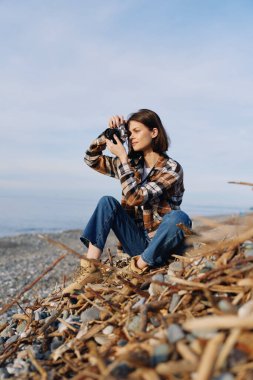 Sahilde fotoğraf makinesi olan sıradan bir fotoğrafçı, okyanus manzarasını yakalıyor, kompozisyon ve ışığa odaklanıyor, saçlarında rüzgar ve sert plaj dokularıyla..