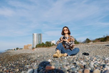 Güneşin tadını çıkaran kayalık bir sahilde uzak şehir silueti ve arkasında sakin bir deniz olan bir kadının olduğu sıradan bir açık hava sahnesi..
