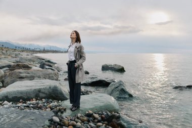 Paltolu sakin kadın okyanusun kenarında bulutlu gökyüzü ile taşların üzerinde duruyor. Kayalar ve suyla doğal manzara sahile yakın huzur ve huzur yansıması yaratır..