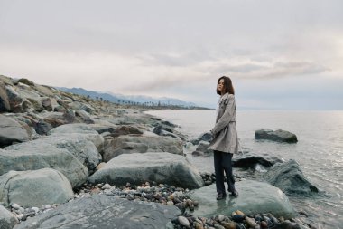 Paltolu kadın bulutlu gökyüzünün altında sakin deniz suyunun yanında kayalık sahilde duruyor. Sahne, sessiz bir atmosferde yalnızlığı ve doğayla bağlantıyı iletir..