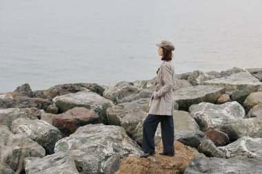 Trençkotlu ve bereli bir kadının sakin denizin kıyısındaki kayalarda duruşu. Bulutlu gökyüzü ve doğal manzara yalnızlığı ve yansımayı vurgular..