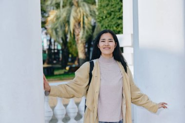 Açık hava sahnesinde, sütunlar, park dekorları, güneş ışığı, esinti, kendine güvenen duruş, günlük zarafet, boş bir ruh hali, yayın tarzı, yaklaşılabilir yaklaşımlar arasında gülümseyen bir kadın var.