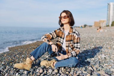 Çakıl taşlı bir sahilde, güneş gözlüğü, ekose gömlek ve kot giyen, kıyı manzarası ve sakin suda ıslanan doğal bir doğa, tatil ve yaşam tarzı imajı olan doğal bir doğa kaşifi.