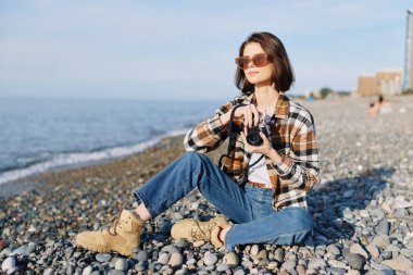 Sıradan bir fotoğrafçı, ekose gömlek ve kot pantolon giyiyor, güneş gözlüğü takıyor, sahilde kameralı oturuyor, saçlarında rüzgar, deniz meltemi, basit deniz manzarası ve yaratıcı yaşam tarzı tasvirleri..