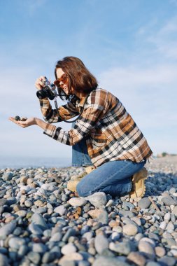 Düz taşların arasında diz çökmüş bir kadın fotoğrafçının olduğu açık hava fotoğrafçılığı sahnesi, temiz bir gökyüzünün altında bir kamerayı hedef alarak, odak, yaratıcılık ve seyahat yaşam tarzını gösteriyor..