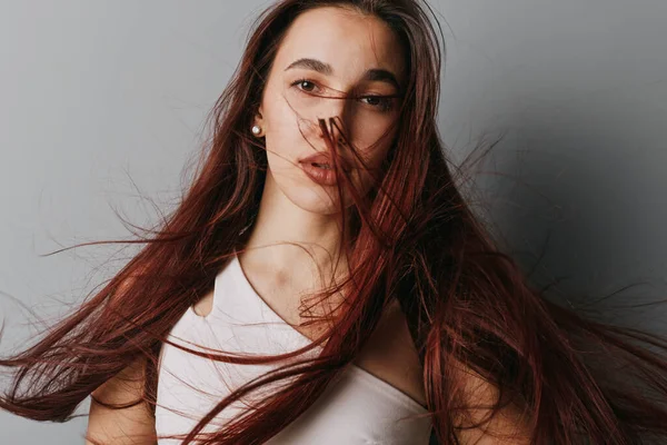 Uzun saçlı, rüzgarda savrulan beyaz bluzlu genç bir kadın. Tarafsız gri arka planda doğal görünümlü ve minimum makyajlı yakın plan portresi..