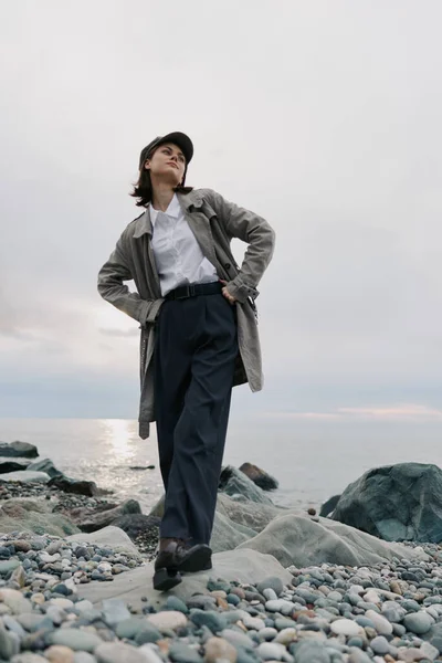 Bulutlu bir günde çakıl taşlı sahilde paltolu ve şapkalı kendine güvenen genç bir kadın poz veriyor. Moda, doğa, açık hava yaşam tarzı, sakin ve doğal ortamda yakalanan sıradan tarz..