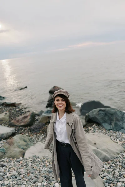 Gündelik giysiler içinde gülümseyen genç bir kadın bulutlu gökyüzünün altında sakin deniz kıyısında duruyor. Şapka takıyor ve uzun bir palto giyiyor. Açık havanın tadını çıkarıyor. Suyun yanında rahat ve mutlu bir havası var..