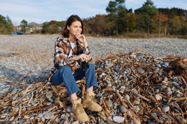 Çakıl taşları ve deniz yosunları, sonbahar ışığı, ekose gömlek ve botları ile sakin bir plajda oturan kadın, taşların arasında oturmuş, rahatlatıcı bir ruh hali ve kırsal kıyı atmosferini yakalıyor.