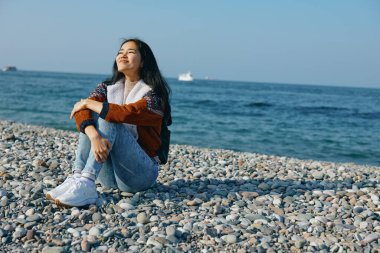 Çakıl taşlı bir sahilde rahat bir kadın, şık bir kıyafet ve sakin bir okyanus arkaplanı, dinlenme, yansıma ve açık hava kaçışını sunar..