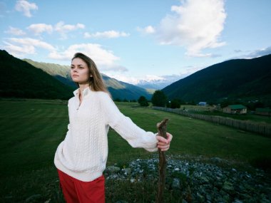 Açık hava sahnesinde örgü kazaklı bir kadın kırsal bir vadide, arka planda dağlar, eldeki personel, parlak gökyüzü ve geniş kırsal alan