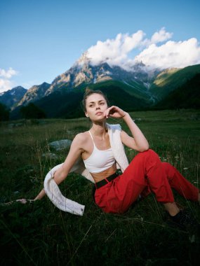 Açık hava manzarasında sıradan bir kadın çimenlerin üzerinde oturuyor, uzaktaki alp dağları, beyaz üst kırmızı pantolon, omuz üstünden atkı, parlak gün ışığı ve sakin doğa manzarası.