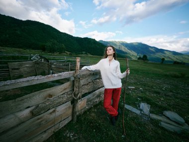 Açık hava sahnesinde beyaz bir kazak ve yıpranmış bir çitin yanındaki kırmızı pantolon, geniş çayır, yuvarlanan tepeler, berrak gökyüzü, kırsal keşif anı...