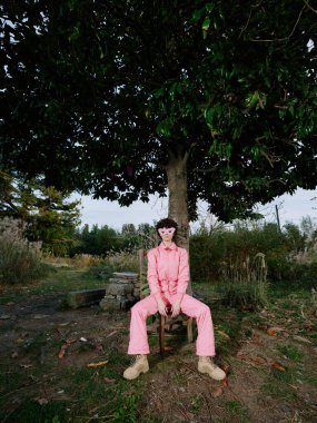 Kırsal bir alanda, yapraklı bir ağacın altında dinlenen pembe tulumlu tek bir figürün portresi. Yumuşak gün ışığı, sakin atmosfer ve toprak bir patikanın yanında rahat duruş ve vahşi.