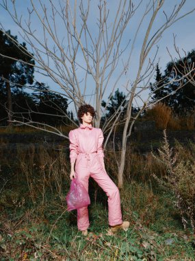 Pembe tulumlu bir mankenin portresi, uzun otlar ve ağaçlar arasında güvenle poz veriyor, sıradan kıyafetleri doğayla harmanlıyor ve şık bir şehir manzarası yaratıyor..