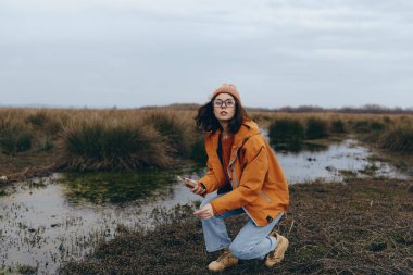 Beyaz kadın gözlük takıyor ve uzun otlarla çevrili ıslak arazide sıcak giysiler giyiyor. Açık hava yaşam tarzı, sonbahar sezonu, doğa yürüyüşü ve günlük moda kavramı..