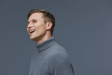 Gri balıkçı yaka kazak giyen gülümseyen beyaz adam gri arka plana karşı mutlu bir ifadeyle bakıyor. Dijital detoks ve zihinsel sağlığı yansıtan günlük yaşam tarzı portresi